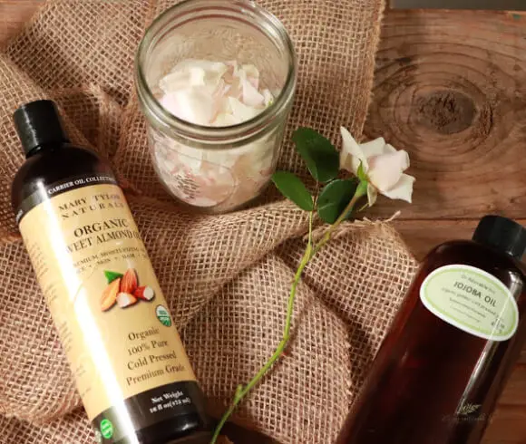 DIY Rose Oil rose petals in mason jar with rosebud alongside as well as sweet almond oil bottle and jojoba oil bottle