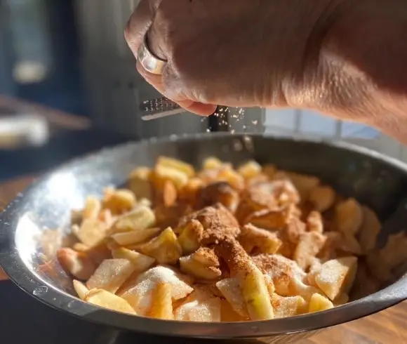 Homemade Gluten-Free Apple Crisp bowl of sliced apples with sugar & cinnamon on top showing a nutmeg being freshly grated over the top