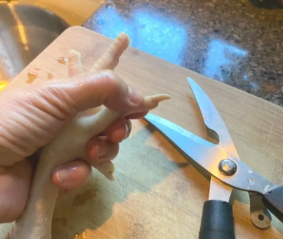 Preparing And Using Chicken Feet hand holding peeled chicken foot over cutting board with a pair of kitchen shears ready to clip the chicken nail off