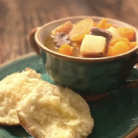 Easy Homemade Beef Stew image showing beef stew in a cup with two handles on a plate with homemade biscuits on the side