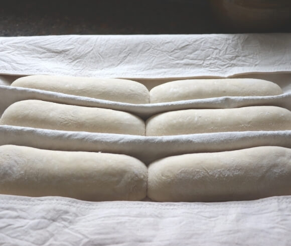 Homemade Baguettes image showing 6 shaped dough rolls resting on floured pastry cloth with bends in the cloth separating the dough rolls