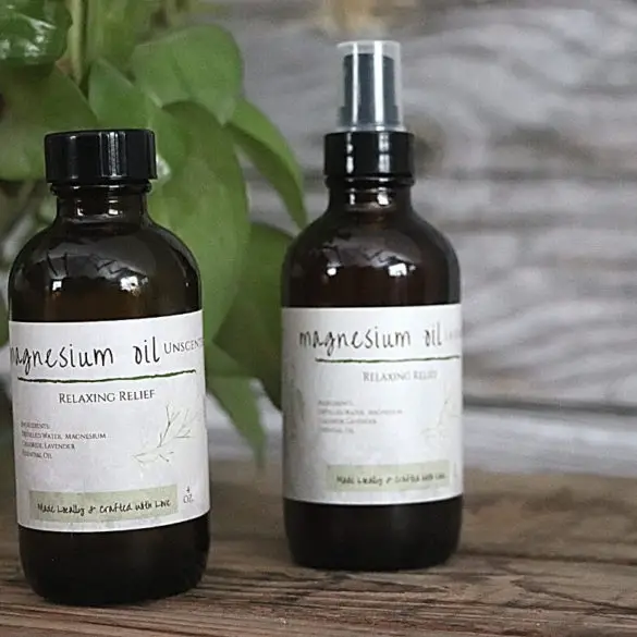 Magnesium Oil Benefits, Uses, And DIY Spray Recipe image showing 2 bottles of magnesium oil spray in front of wooden board and greenery