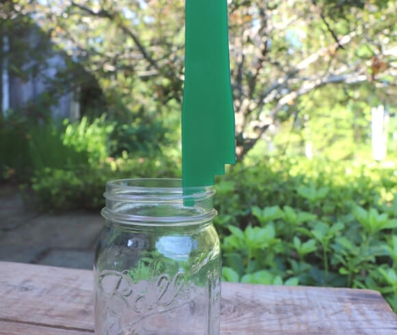Headspace For Canning image showing quart size mason jar resting on wooden board with blurred greenery in background displaying a green headspace measurement tool resting on lip of jar