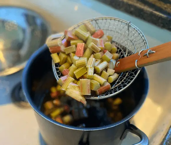 How to Freeze Rhubarb image showing cut up rhubarb pieces being lifted out of a blanching bath of hot water