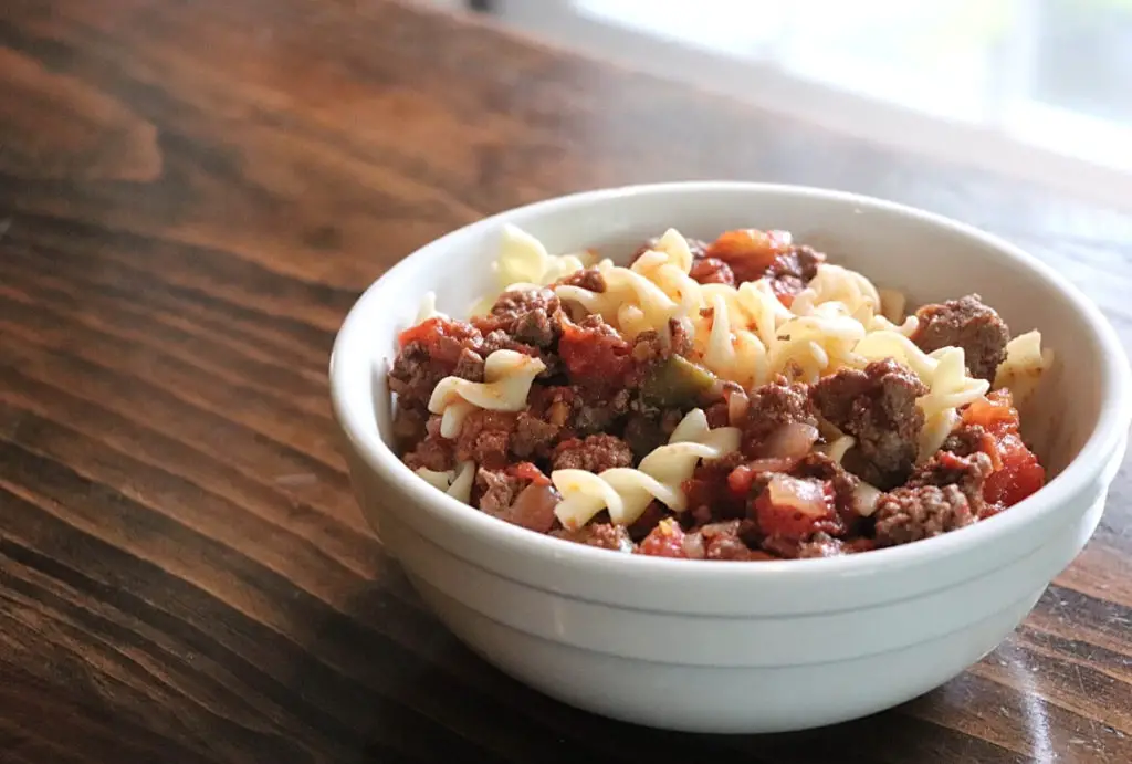 New England Style American Chop Suey featured image showing a white bowl of goulash or American chop suey resting on a wooden board top