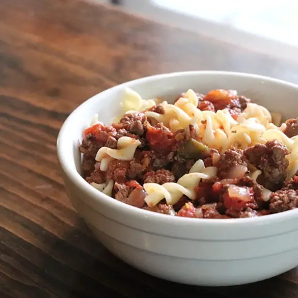 New England Style American Chop Suey featured image showing a white bowl of goulash or American chop suey resting on a wooden board top