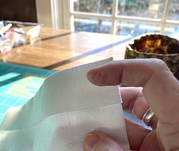 DIY Linen Bread Bag image showing hand holding white linen by sewing table pointing to a one inch edge which has been turned over at the top of the material
