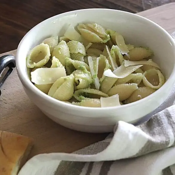 Garlic Scape Pesto Recipe image showing bowl with shell shaped pasta and garlic scape pesto resting on a wooden board with a striped brown and white towel laying next to the bowl