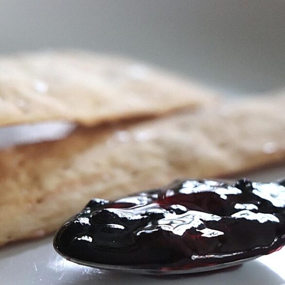 Elderberry Jam Recipe featured image showing spoon filled with deep burgundy colored elderberry jam with several cracker blurred in the background