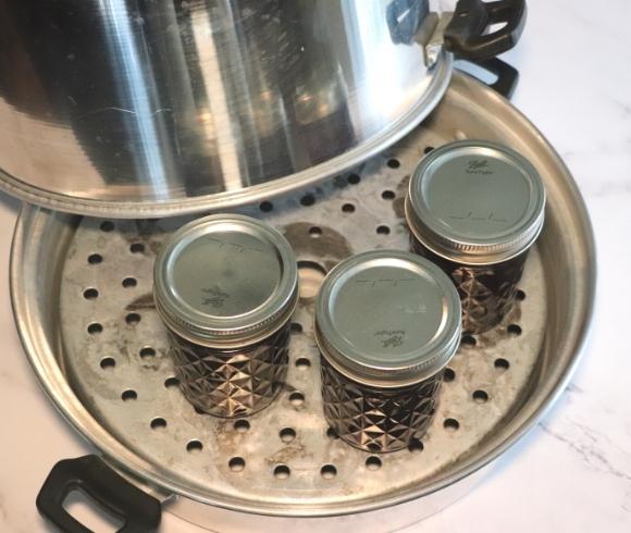 Steam Canning A Complete Guide image showing a top view of 3 jars of elderberry jam resting on the base and rack of a steam canner with the domed steam canning lid resting on the side of the base 