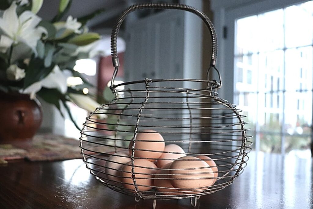 How To Test Eggs For Freshness {Egg Test For Freshness} featured image showing wire egg basket holding fresh eggs resting on a wooden table top with blurred background