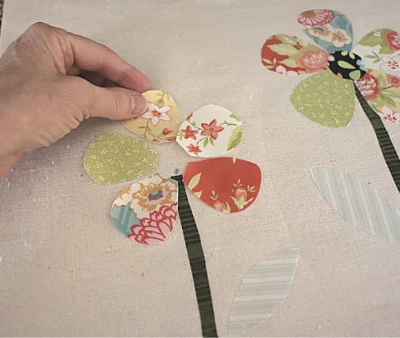 How To Appliqué image showing hand placing cutout motifs to be pressed onto cream colored fabric creating a 5 petaled flower