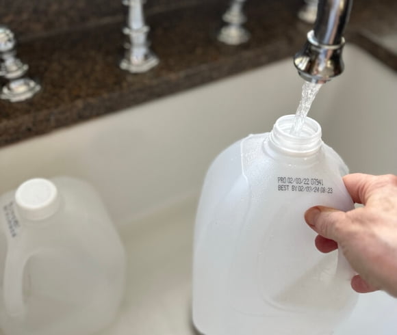 Winter Sowing In Milk Jugs image showing hand holding an open milk jug with water pouring into it from faucet